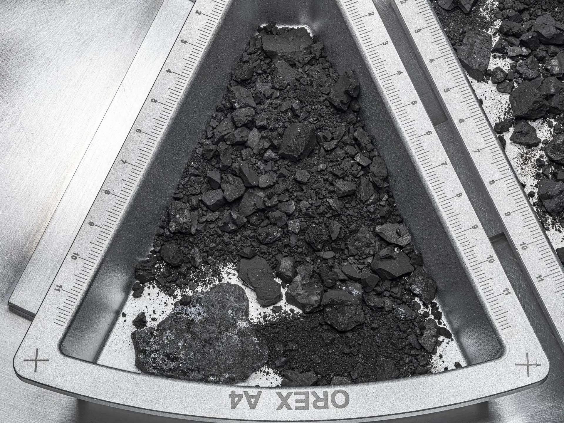 Uma bandeja metálica triangular com rochas pretas e poeira do asteroide Bennu, com réguas em centímetros nas bordas
